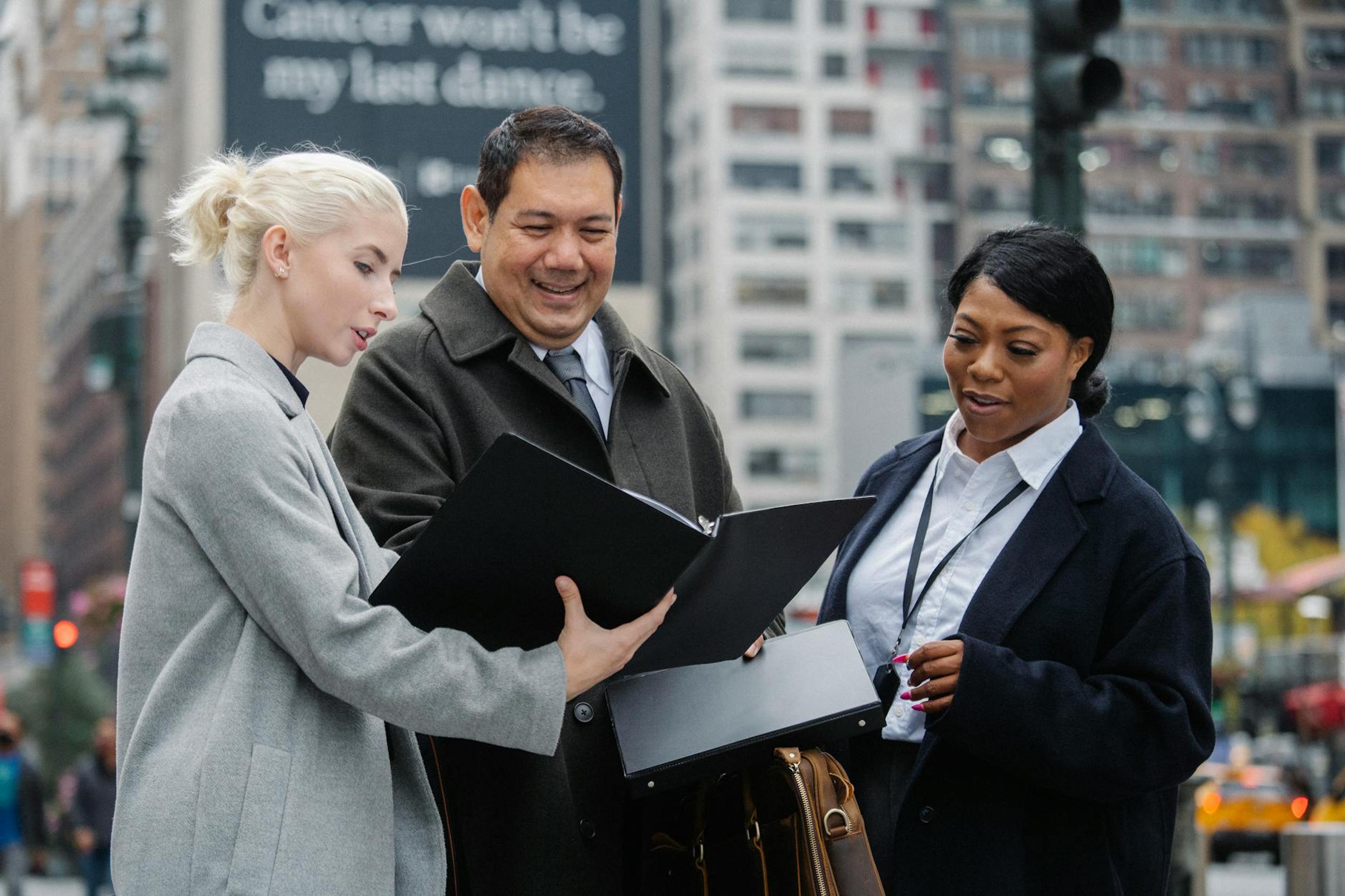 Young well dressed woman sharing folder with multiracial colleagues while talking in town in daytime