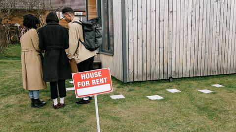 Group of adults viewing a wooden house with a 'House for Rent' sign on the lawn.