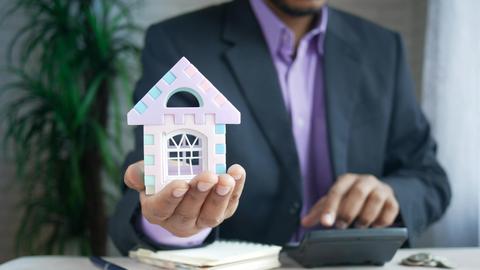 Businessman in suit holds model house, calculates real estate investments.