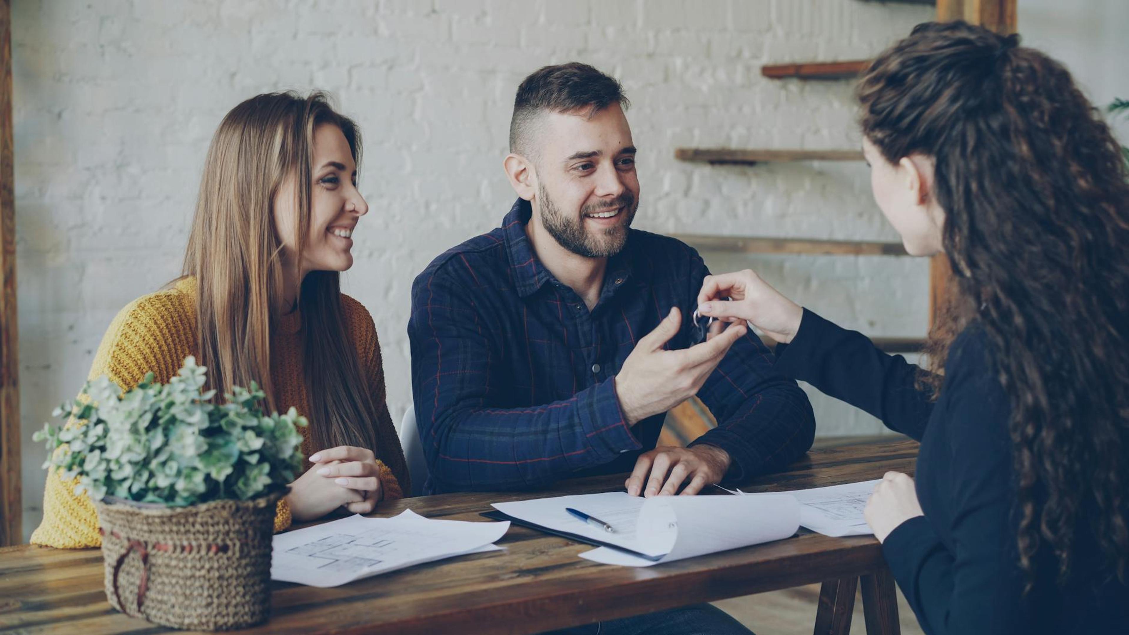 Smiling couple receiving house keys from a female real estate agent at a wooden desk.