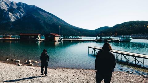 Two people walk towards a lake with mountains.