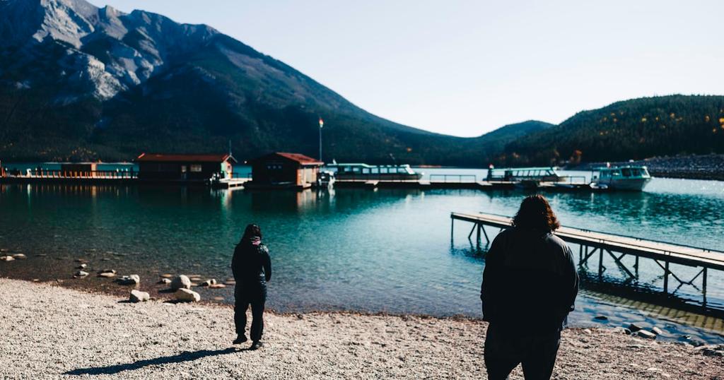 Two people walk towards a lake with mountains.