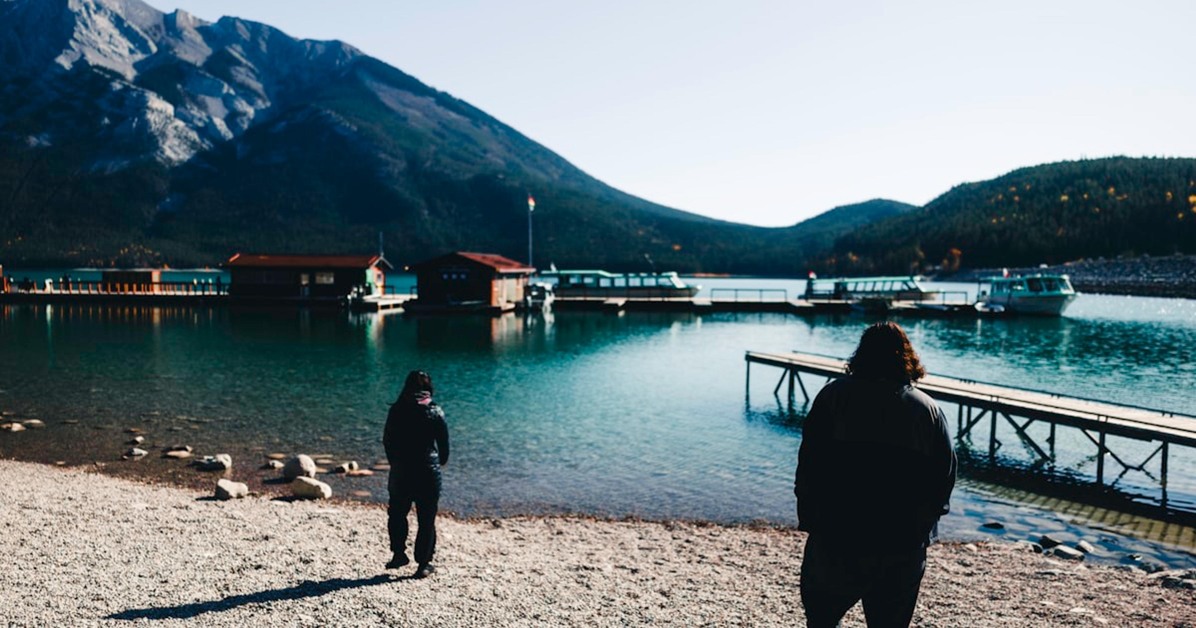 Two people walk towards a lake with mountains.