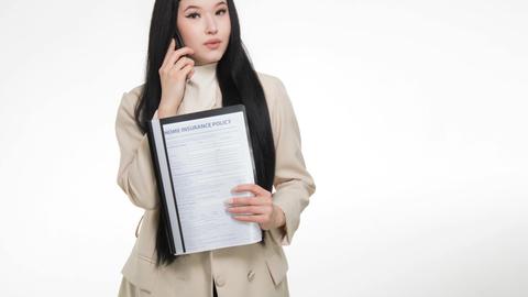 Asian woman in business attire holding home insurance policy while on a phone call.