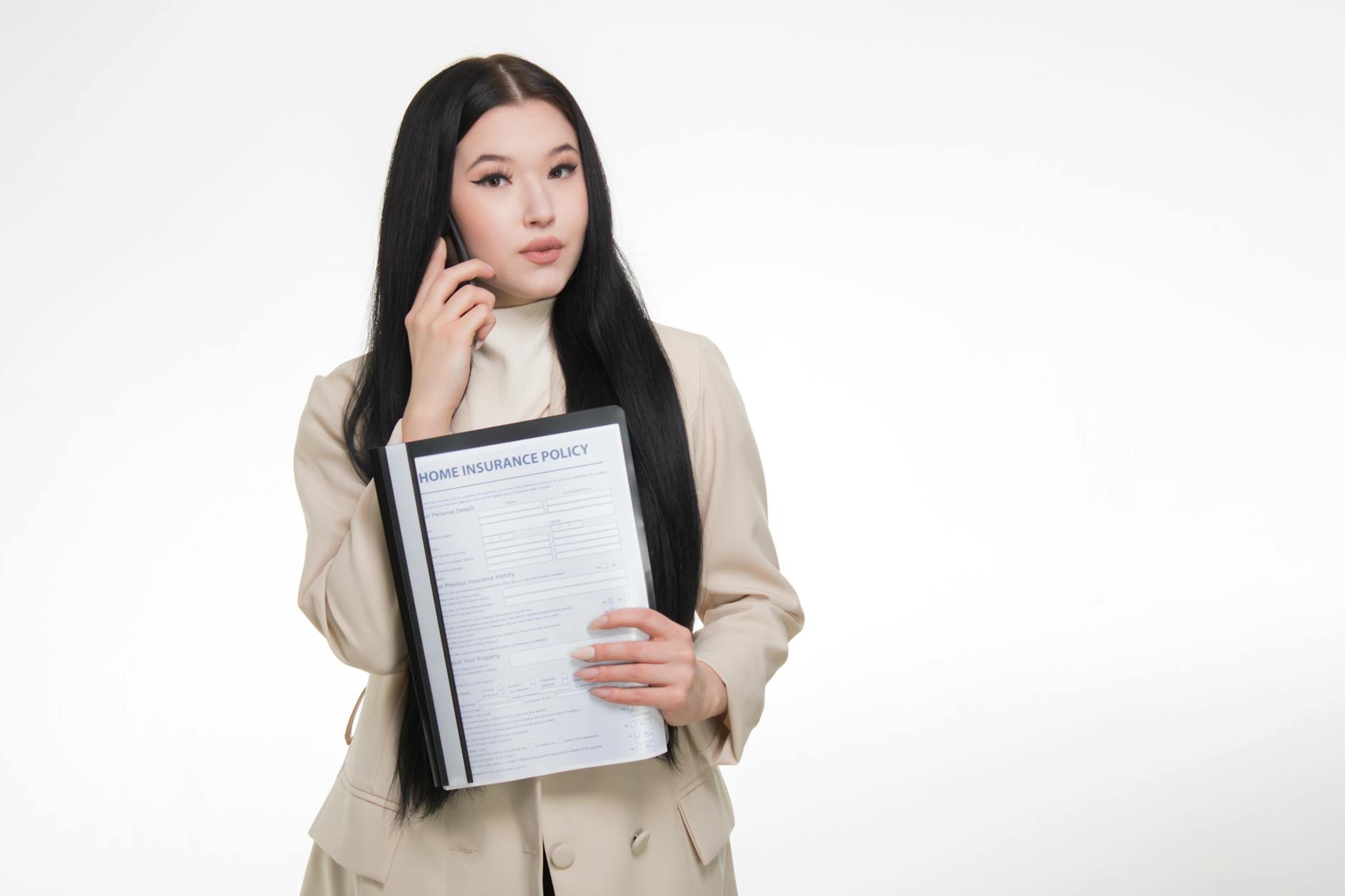 Asian woman in business attire holding home insurance policy while on a phone call.