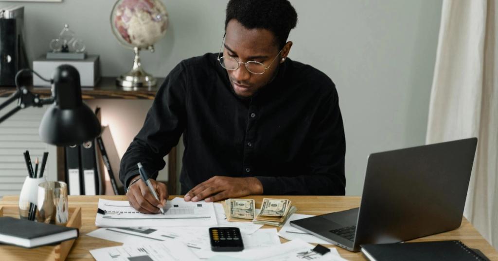 An adult man calculates expenses, using a laptop and documents at a desk in a home office setting.