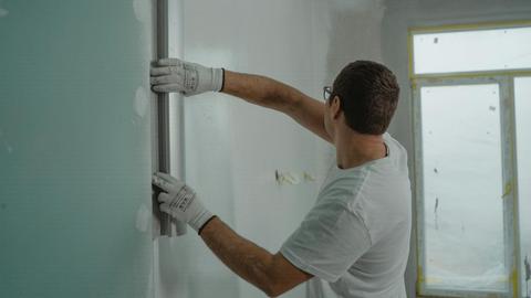 A man in a white shirt and gloves using a metal ruler for indoor wall renovation work.
