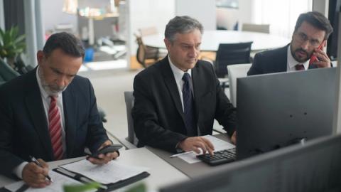Three businessmen in suits working on computers and documents in a modern office setting.