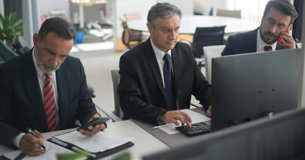 Three businessmen in suits working on computers and documents in a modern office setting.