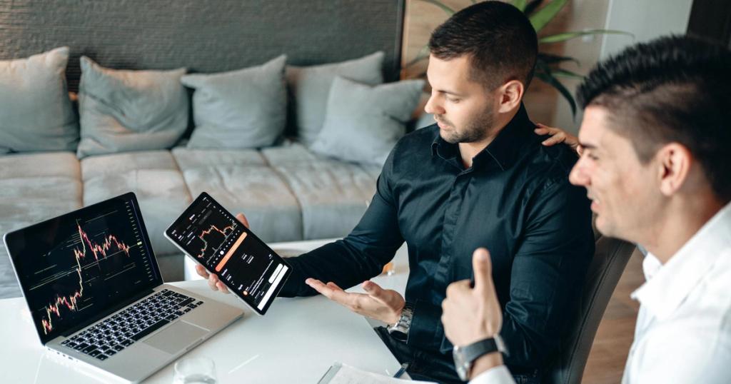 Two businessmen discuss digital trading insights with laptop and tablet in a modern office setting.
