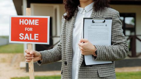 A real estate agent holding a home for sale sign and clipboard outside a property.