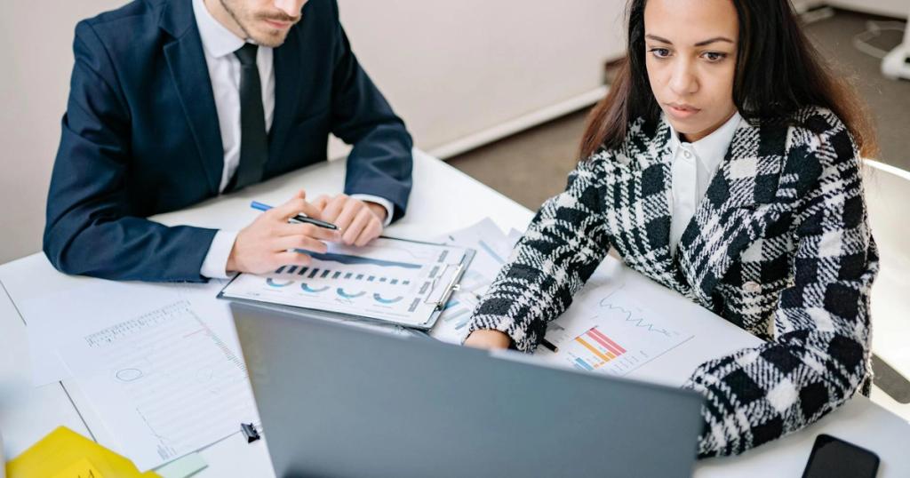 A man and woman collaborate on business analysis at a desk with charts in an office setting.