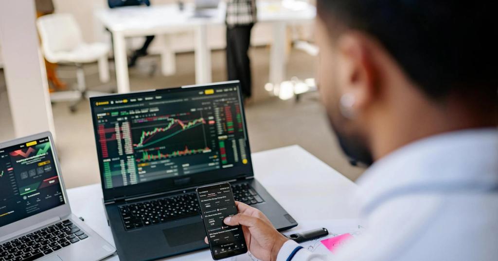 A businessman using smartphone and laptop to track stock market trends in a modern office setting.