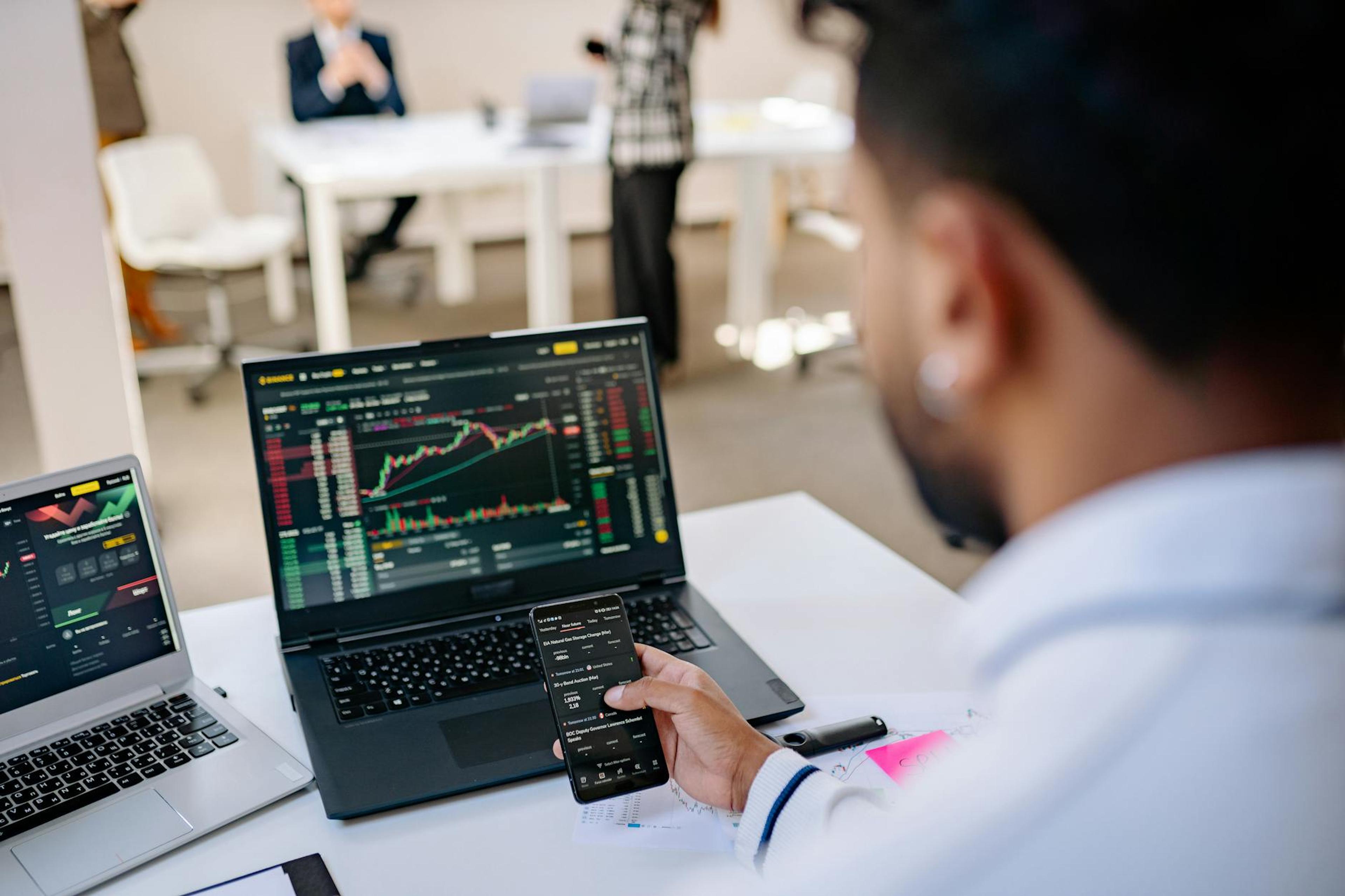 A businessman using smartphone and laptop to track stock market trends in a modern office setting.
