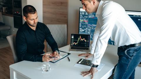 Two businessmen discussing digital market trends with laptops and tablets in a modern office.