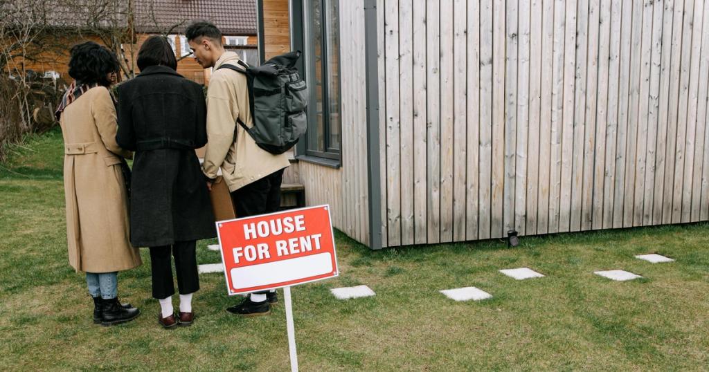 Group of adults viewing a wooden house with a 'House for Rent' sign on the lawn.