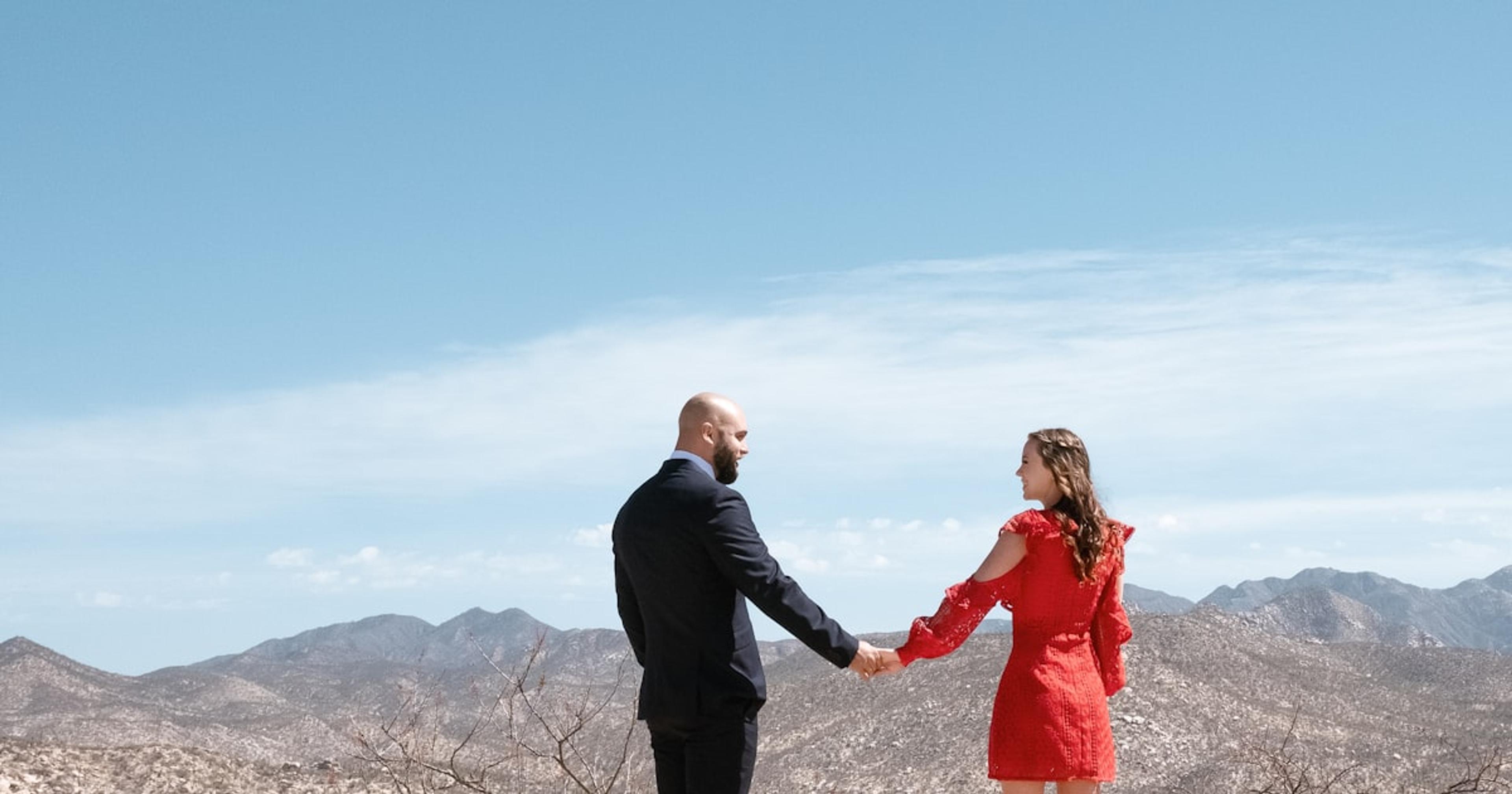 man in black suit jacket holding woman in red dress on top of mountain during daytime