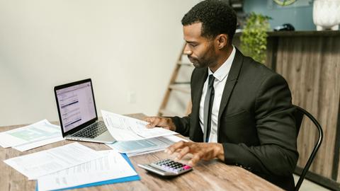 A businessman in a suit working on documents and using a laptop at an office desk.
