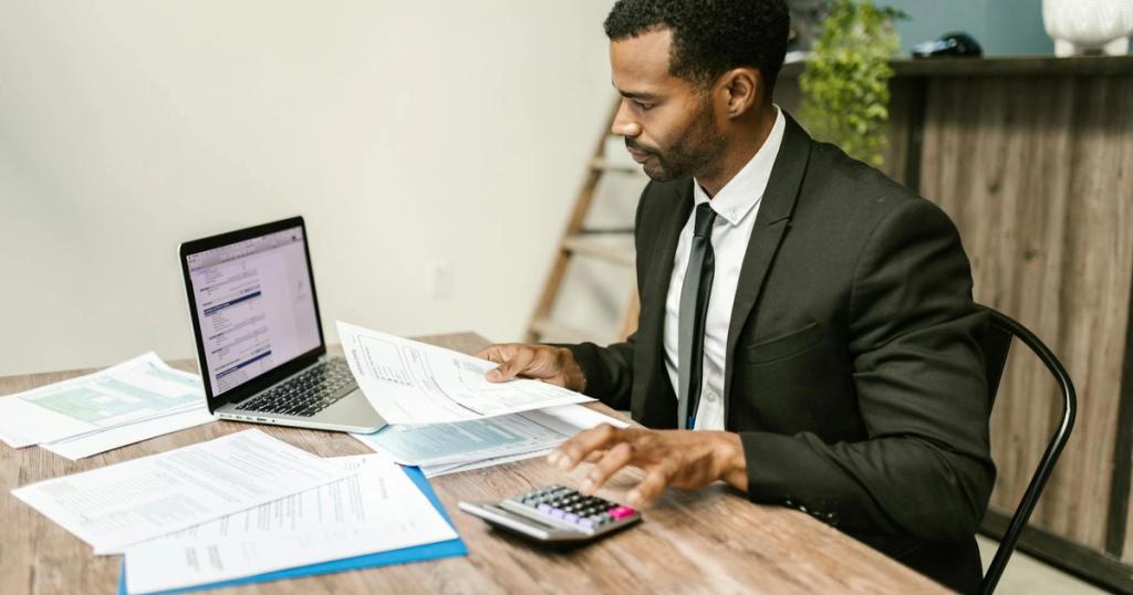 A businessman in a suit working on documents and using a laptop at an office desk.