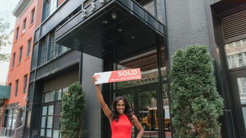 Woman in red dress holds 'SOLD' sign outside office building, symbolizing real estate success.
