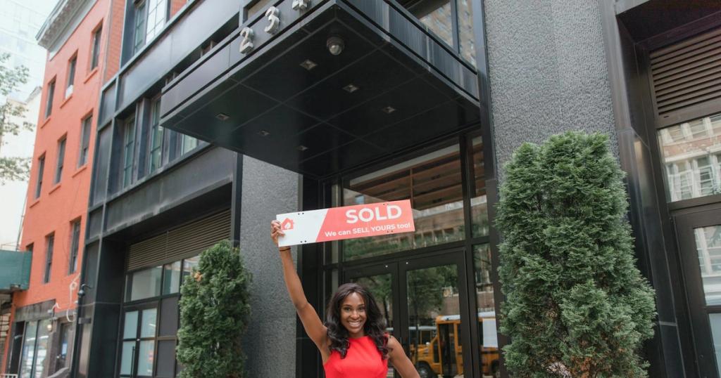 Woman in red dress holds 'SOLD' sign outside office building, symbolizing real estate success.