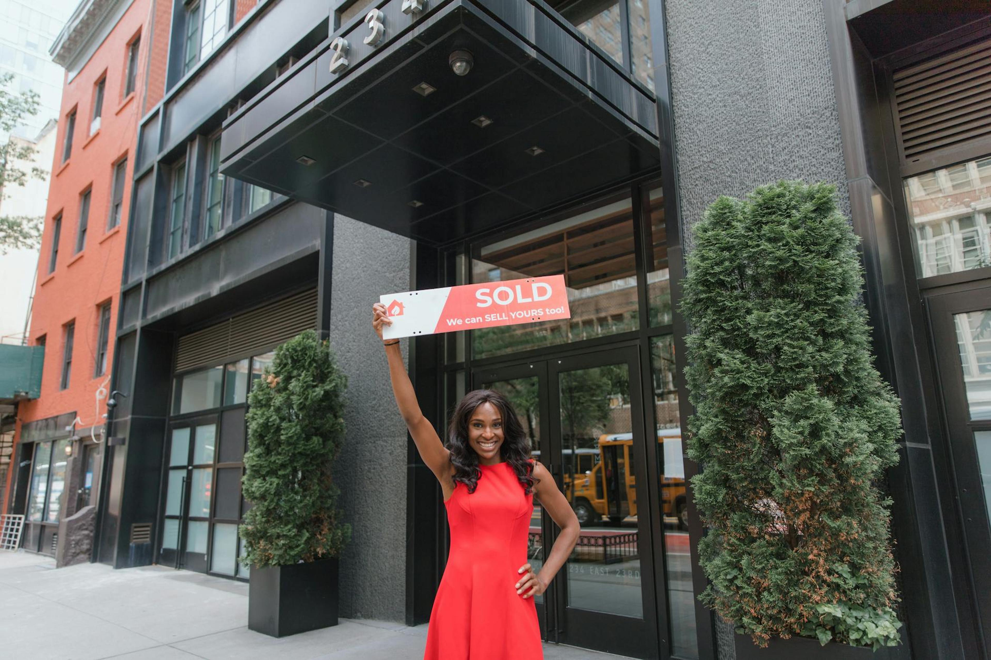 Woman in red dress holds 'SOLD' sign outside office building, symbolizing real estate success.