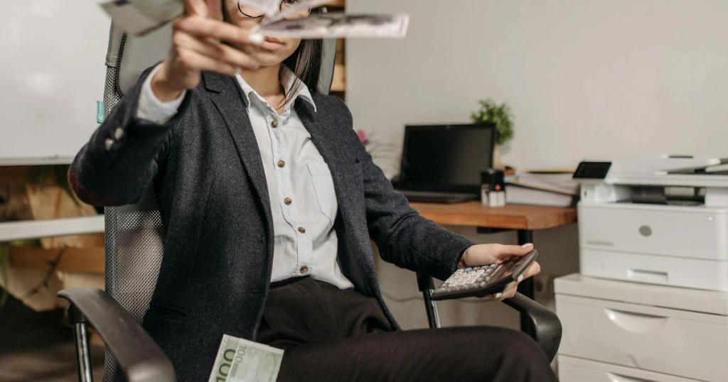 A businesswoman in a suit throws money while calculating finances in a modern office.