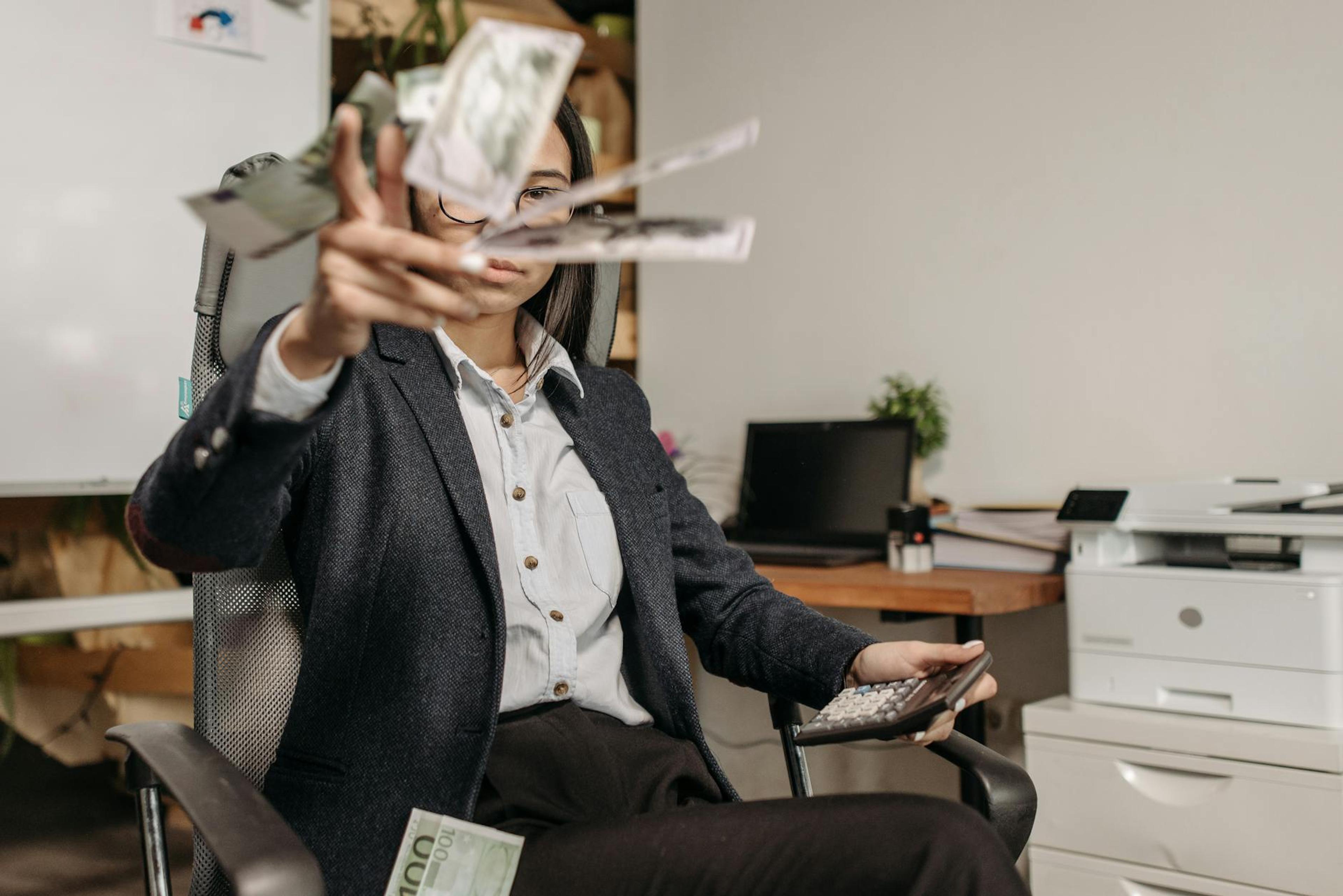 A businesswoman in a suit throws money while calculating finances in a modern office.