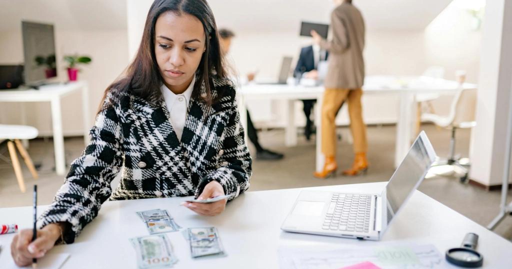 Businesswoman calculating finances with cash and a laptop in a modern office.