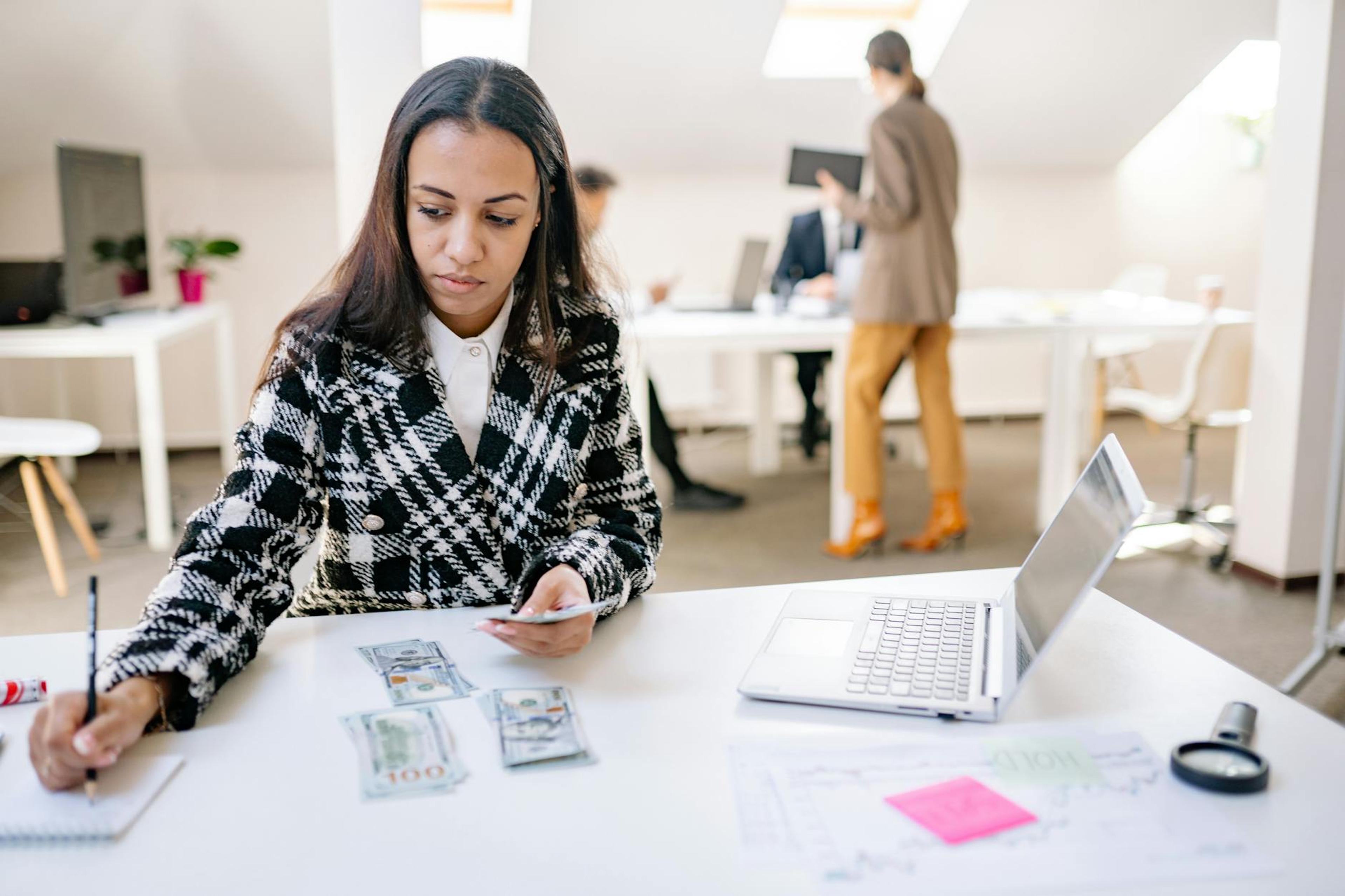 Businesswoman calculating finances with cash and a laptop in a modern office.