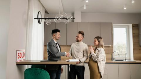A smiling couple and a realtor finalize a house purchase in a modern kitchen setting, sealing the deal with a handshake.