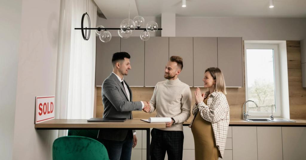 A smiling couple and a realtor finalize a house purchase in a modern kitchen setting, sealing the deal with a handshake.
