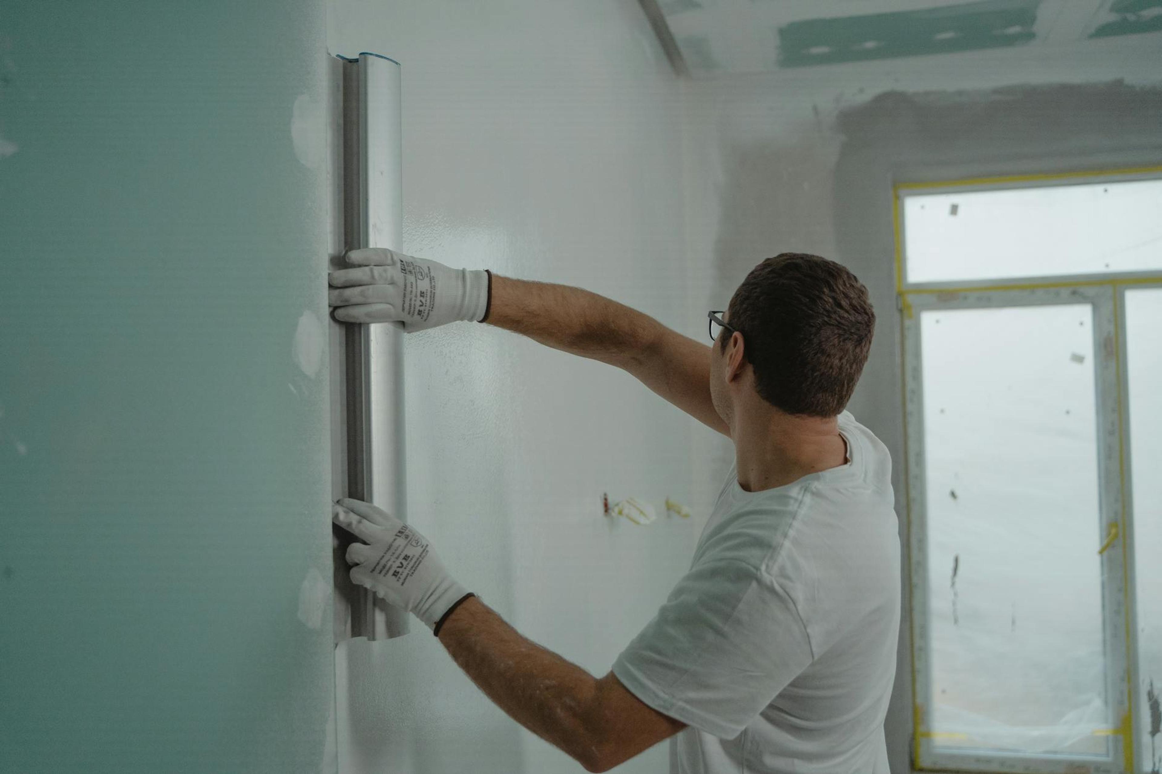A man in a white shirt and gloves using a metal ruler for indoor wall renovation work.