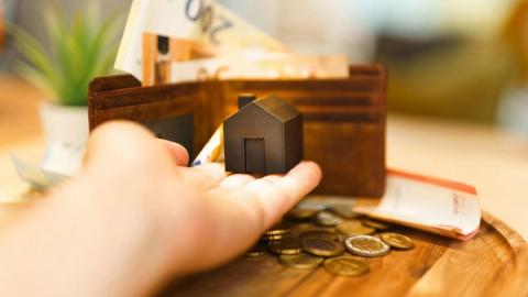 A hand holding a small house model with euro notes and coins nearby, illustrating real estate investment and finance.