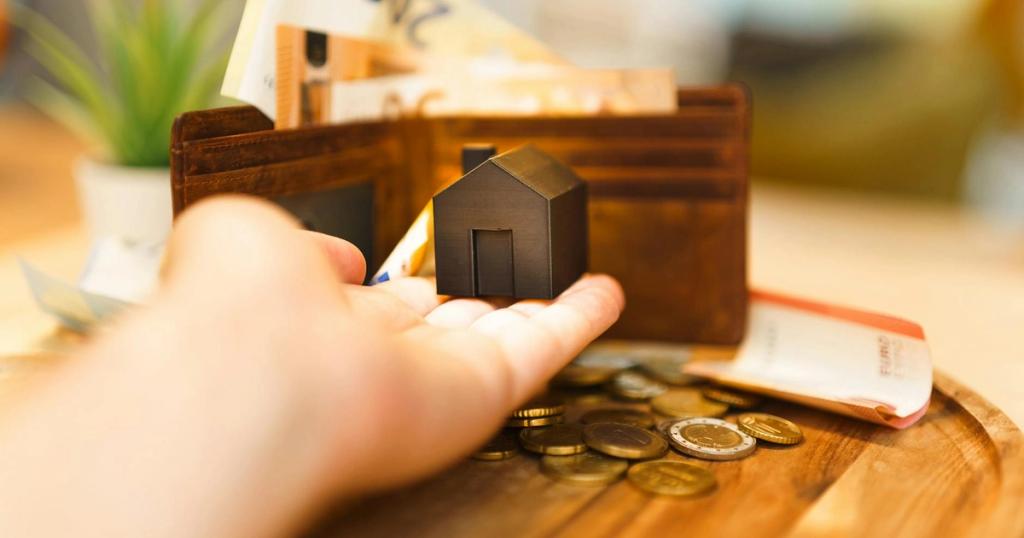 A hand holding a small house model with euro notes and coins nearby, illustrating real estate investment and finance.