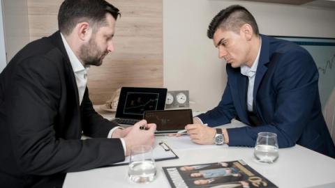 Two businessmen in suits discussing cryptocurrency trends using digital devices in an office setting.