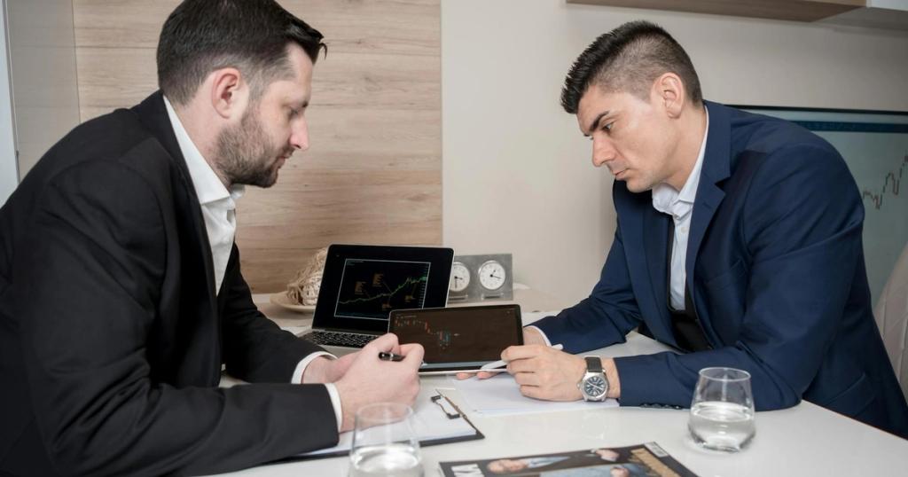 Two businessmen in suits discussing cryptocurrency trends using digital devices in an office setting.