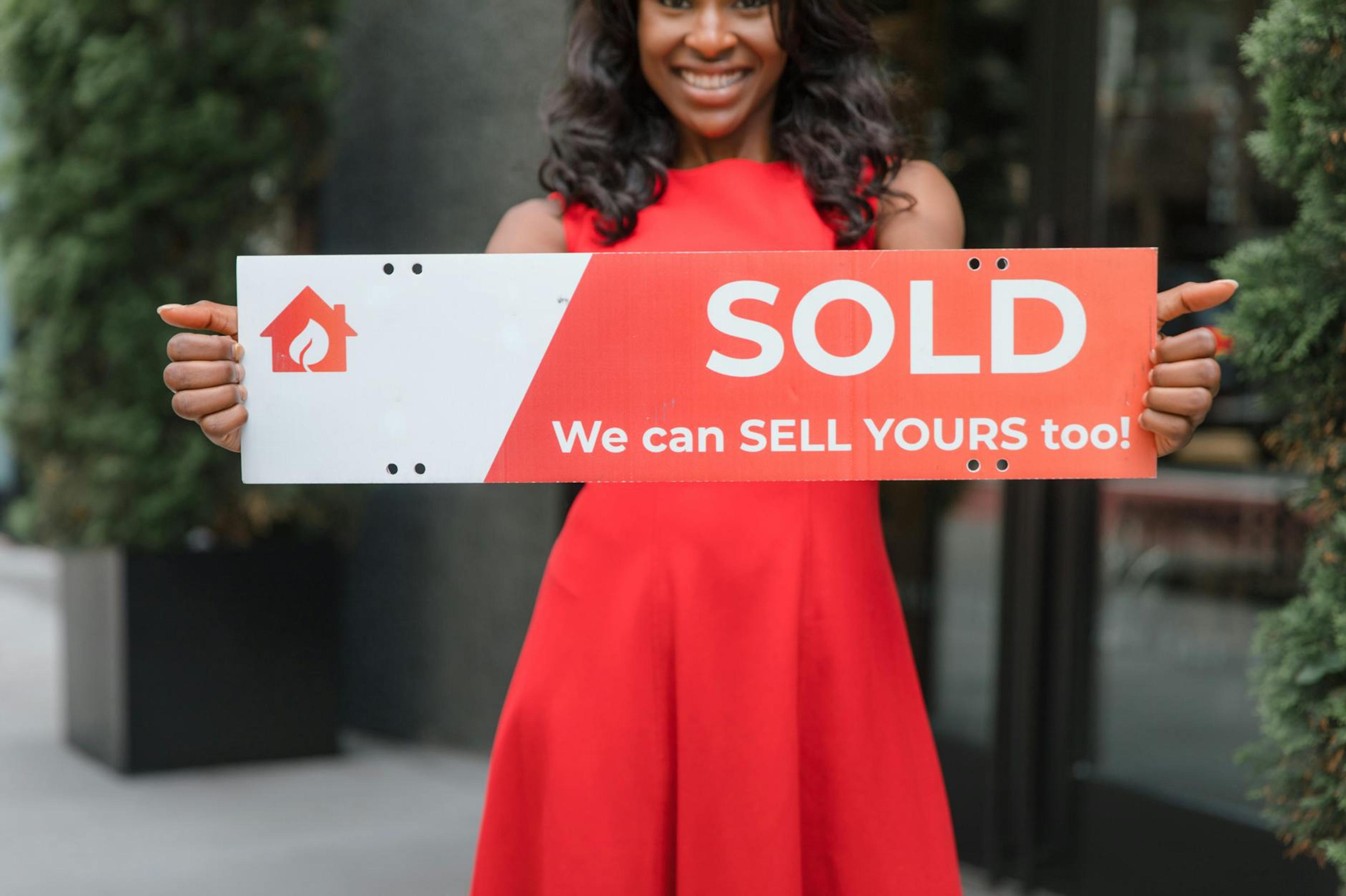 Smiling woman holding a 'Sold' sign, symbolizing successful real estate sales.