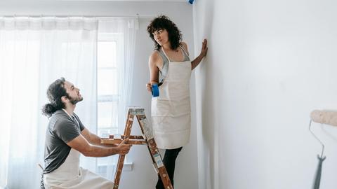 A couple in aprons working together on room renovation using paint rollers indoors.