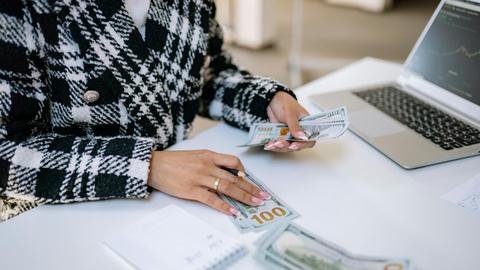Close-up of a woman counting hundred-dollar bills at a desk with a laptop, focusing on finance and work.