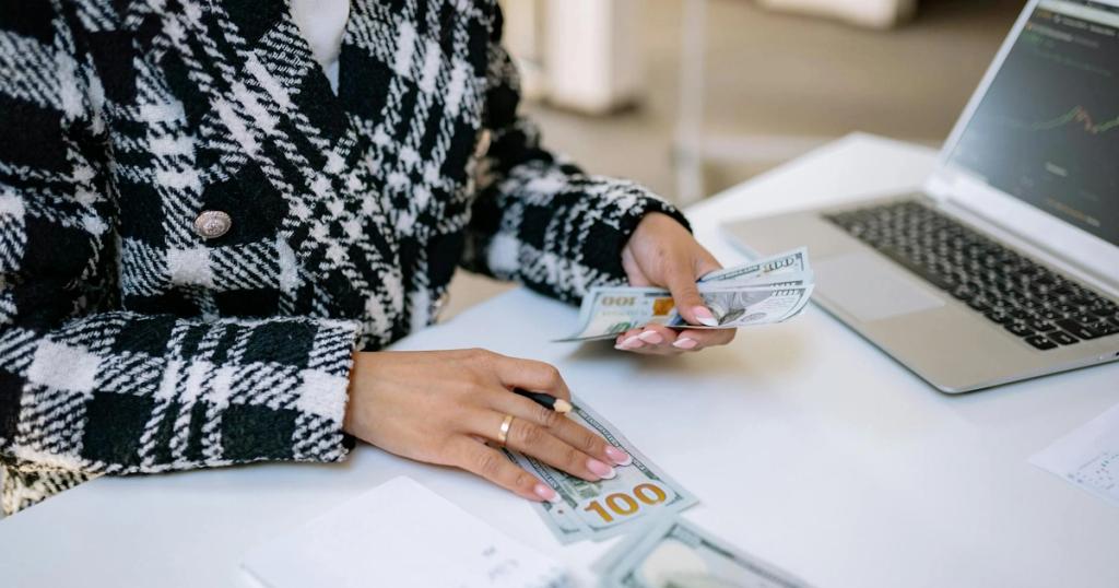 Close-up of a woman counting hundred-dollar bills at a desk with a laptop, focusing on finance and work.