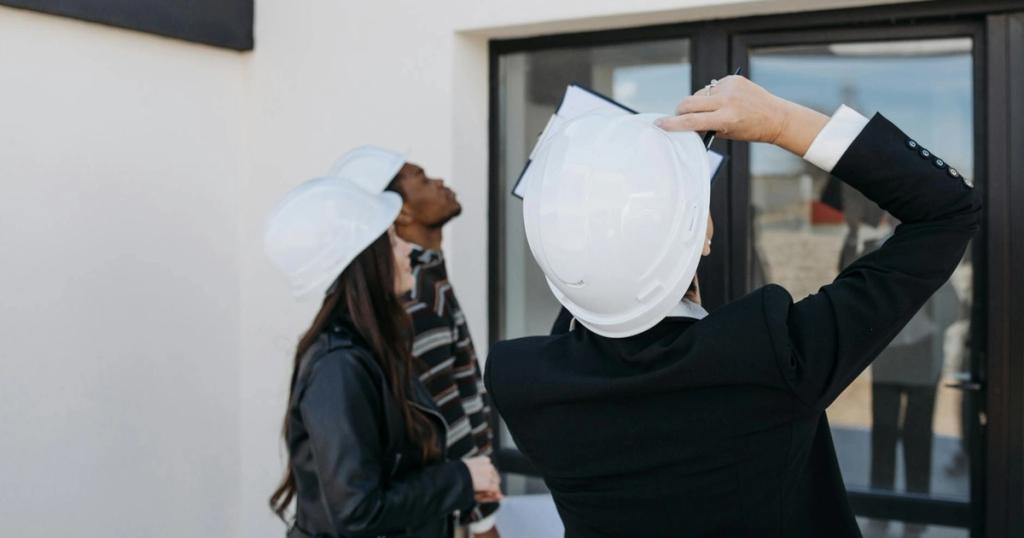Real estate agents wearing hard hats inspect a property's exterior, assessing potential investments.