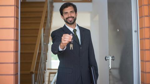 Smiling realtor in a black suit handing keys through an open door inside a modern house.