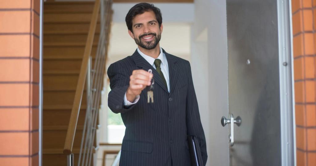 Smiling realtor in a black suit handing keys through an open door inside a modern house.