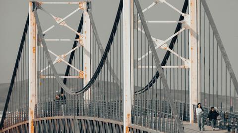 Suspension bridge with visible rust and people walking along, showcasing architecture.