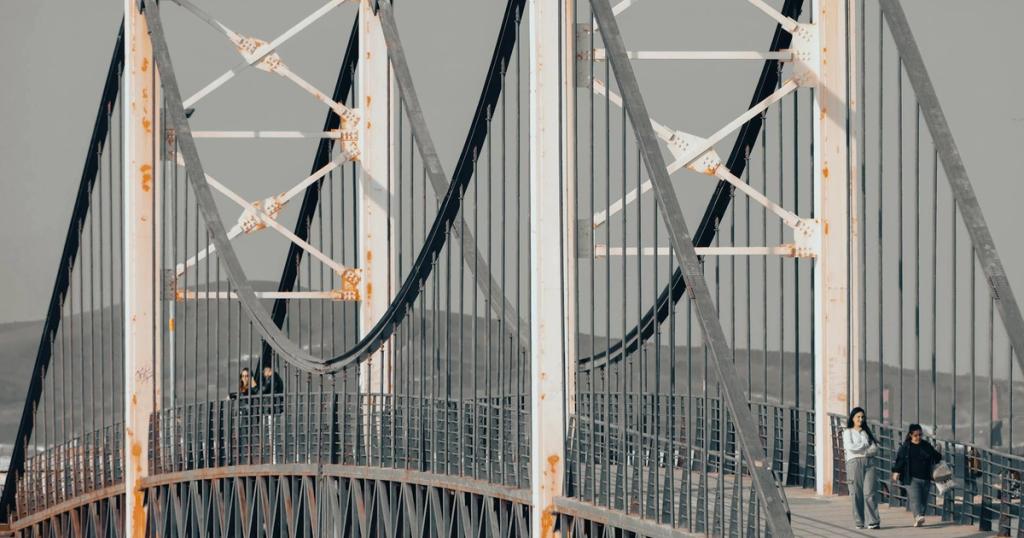 Suspension bridge with visible rust and people walking along, showcasing architecture.