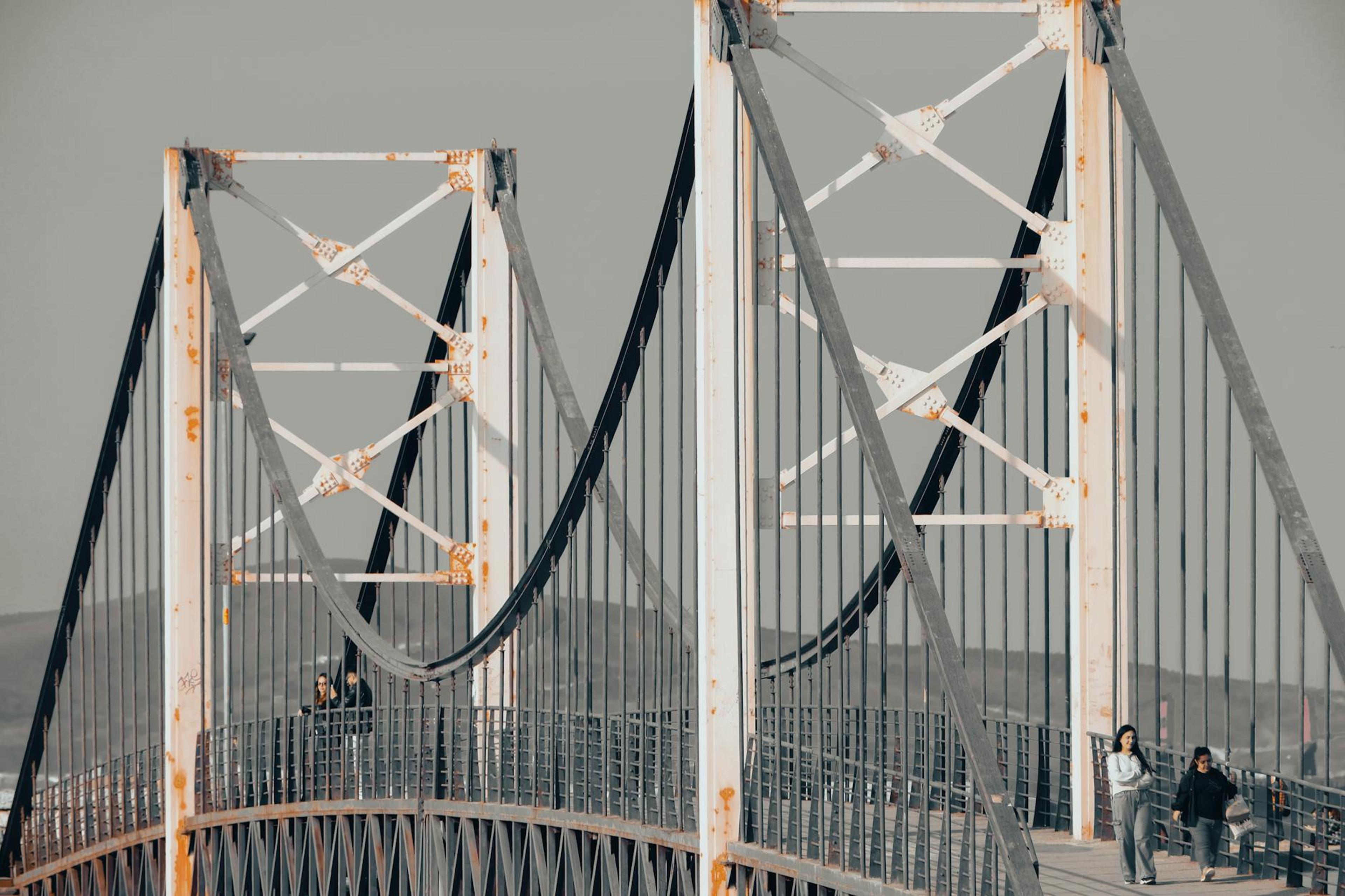 Suspension bridge with visible rust and people walking along, showcasing architecture.