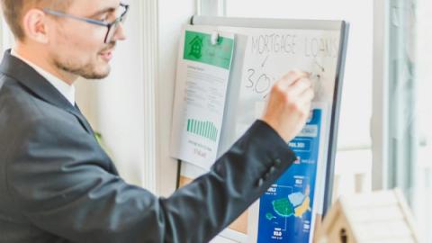 Businessman in suit writes on whiteboard displaying mortgage loan rates during a real estate meeting.
