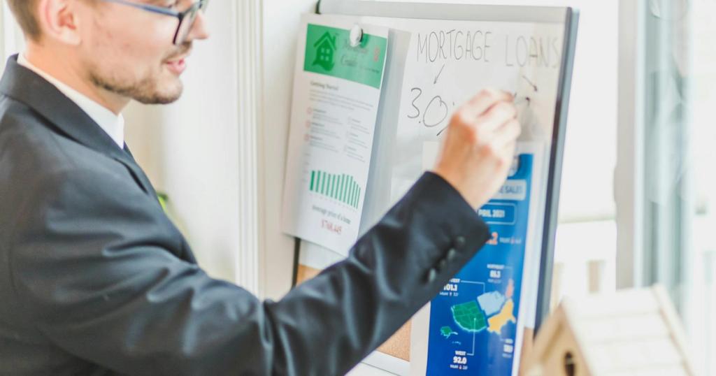 Businessman in suit writes on whiteboard displaying mortgage loan rates during a real estate meeting.