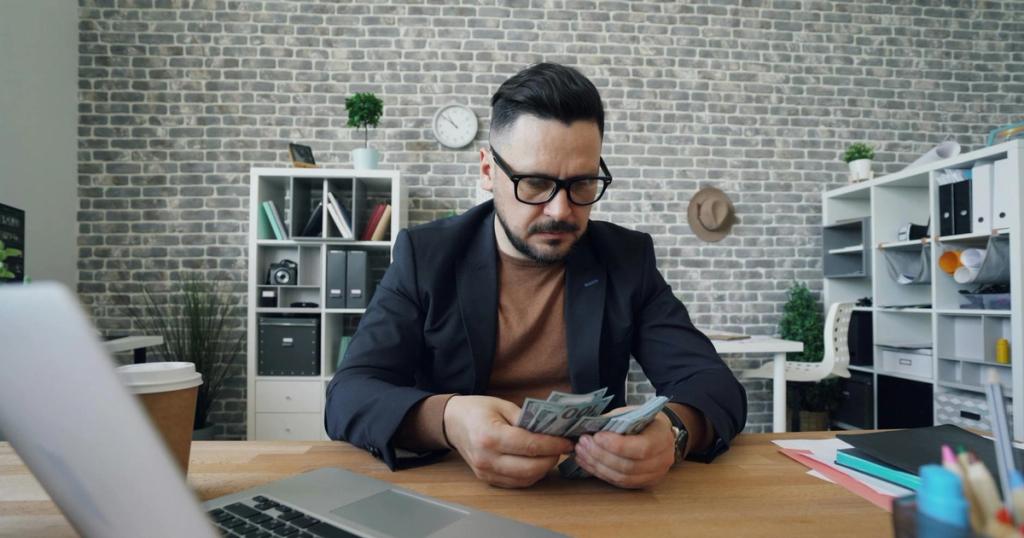 Businessman counting cash at a desk in a modern office setting with a laptop and documents.